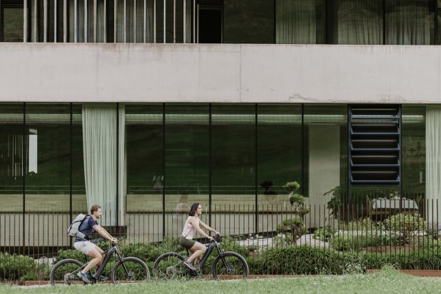 Paar auf E-Mountainbikes fährt an der Glas-Beton-Fassade mit Garten vorbei