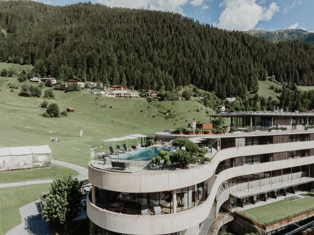 The curved building with Rooftop Pool, with the golf course meadow and wooded mountainside behind