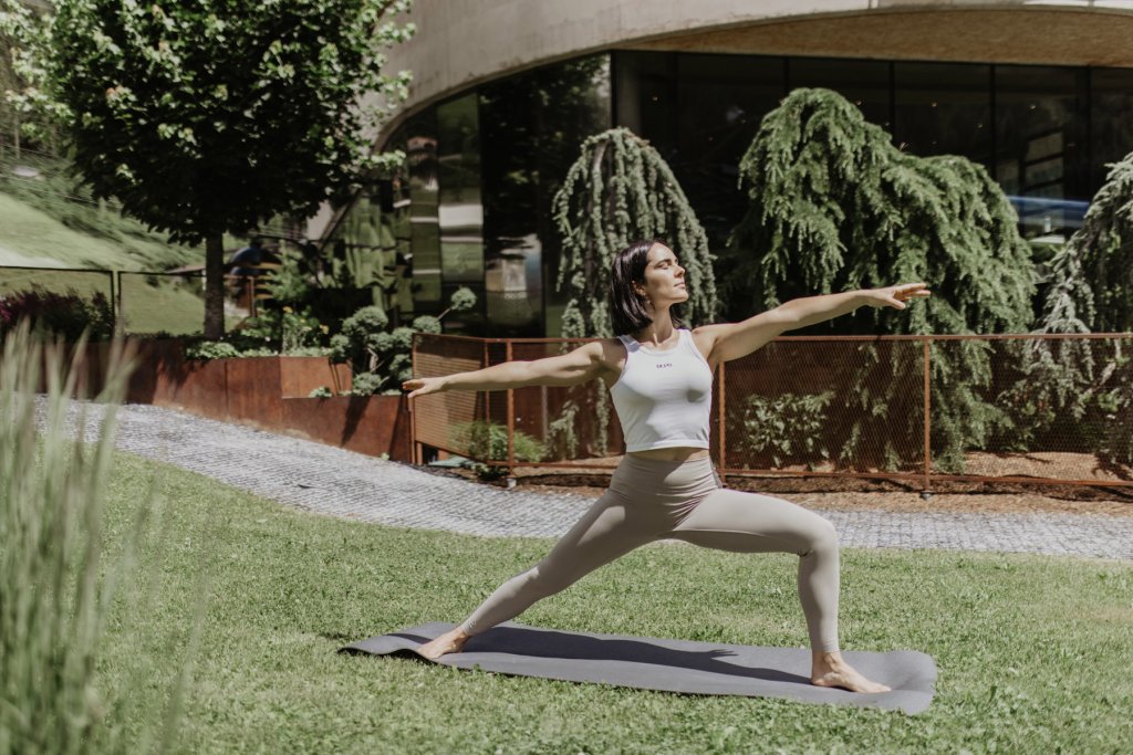Frau in der Krieger-Pose auf der Yogamatte auf der Wiese vor dem Gebäude