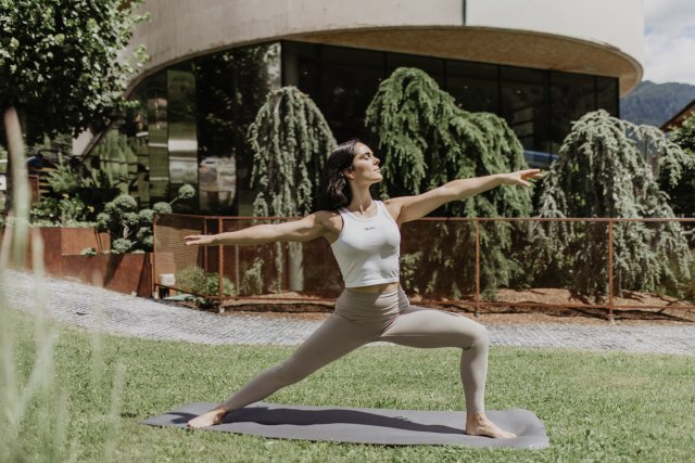 Frau übt Yoga auf der Wiese, dahinter das Gebäude und ein Berghang