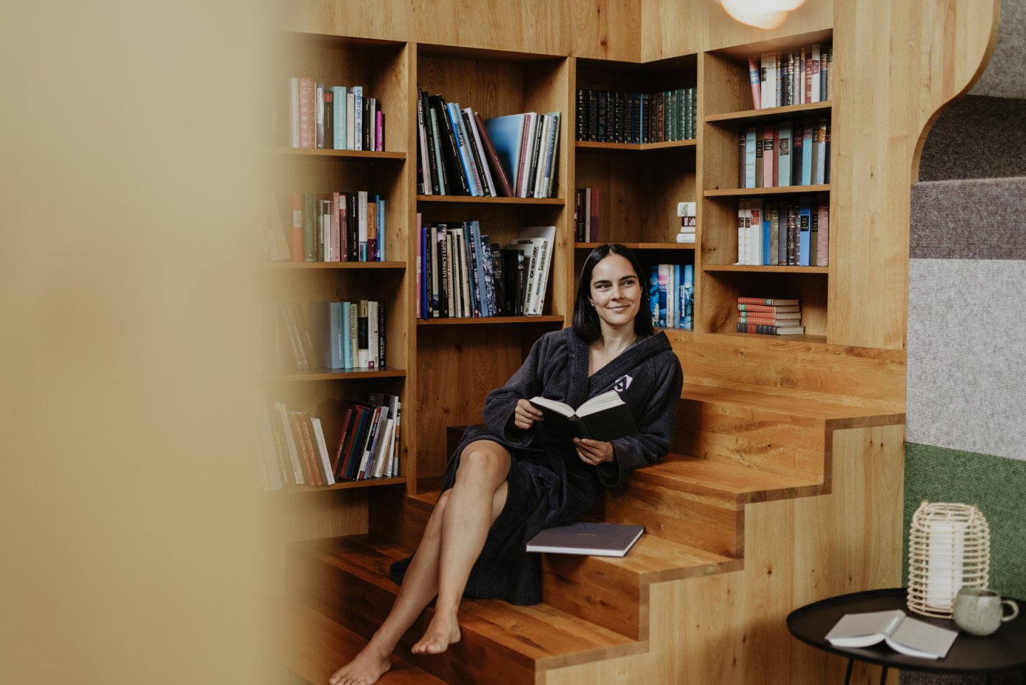 Frau im Bademantel liest auf den Holzstufen vor der Bücherwand