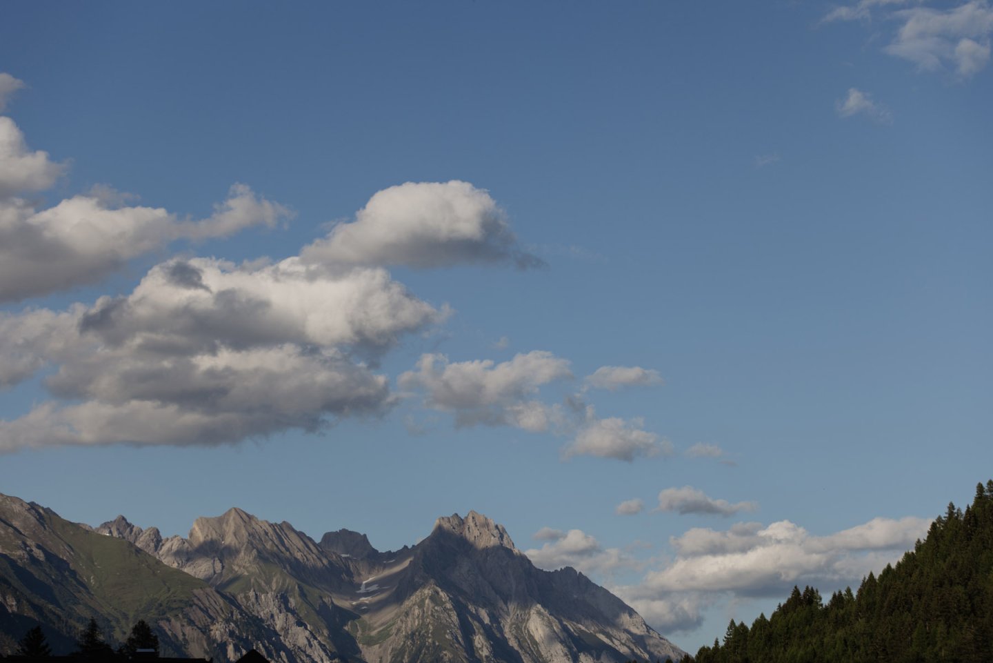Zerklüftete Felsgipfel mit Wolken über bewaldeten Hängen bei blauem Himmel