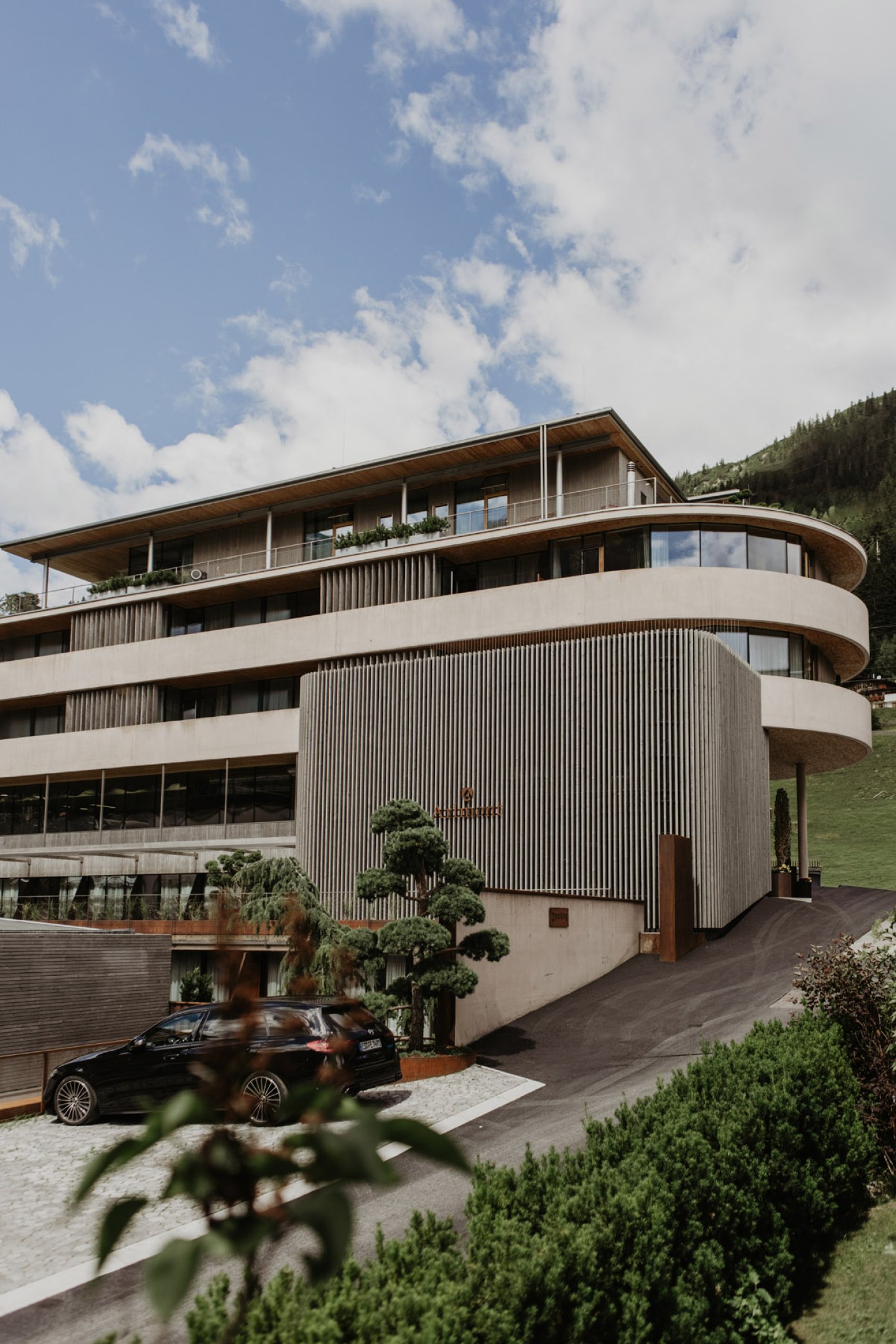 Das geschwungene Gebäude aus Sichtbeton und Holzlamellen mit Zufahrt und geschnittenen Formgehölzen