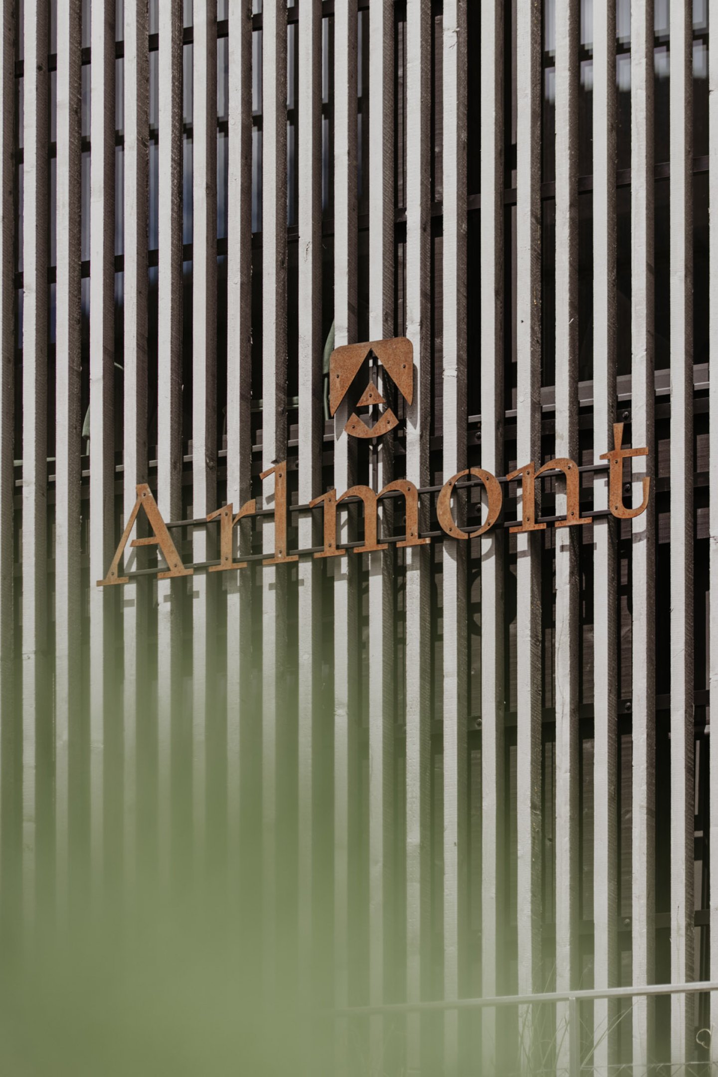 Der Arlmont-Schriftzug aus Cortenstahl an der Holzlamellen-Fassade, darunter eine grüne Spiegelung