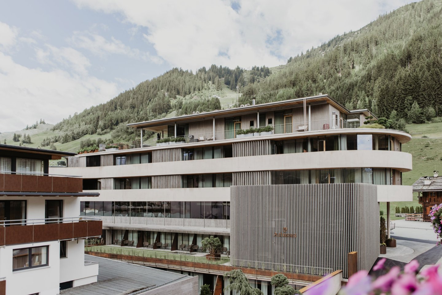 The building from the side with its curved architecture, with a wooded mountainside and the village behind