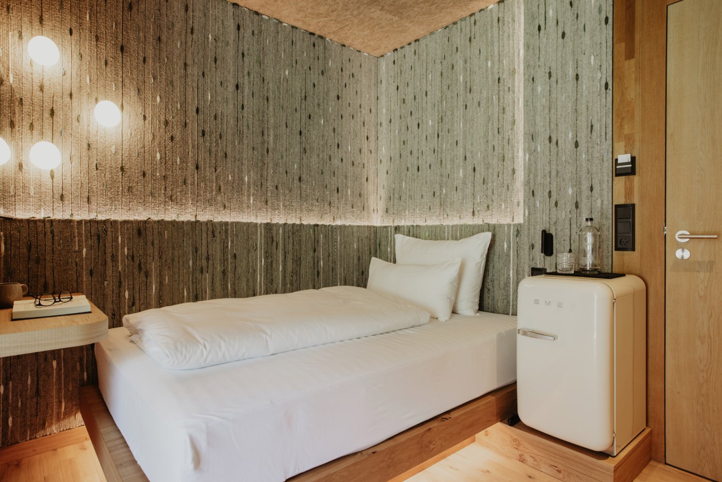 Single bed with white linen against the woven wall panelling, beside it a cream-coloured SMEG minibar