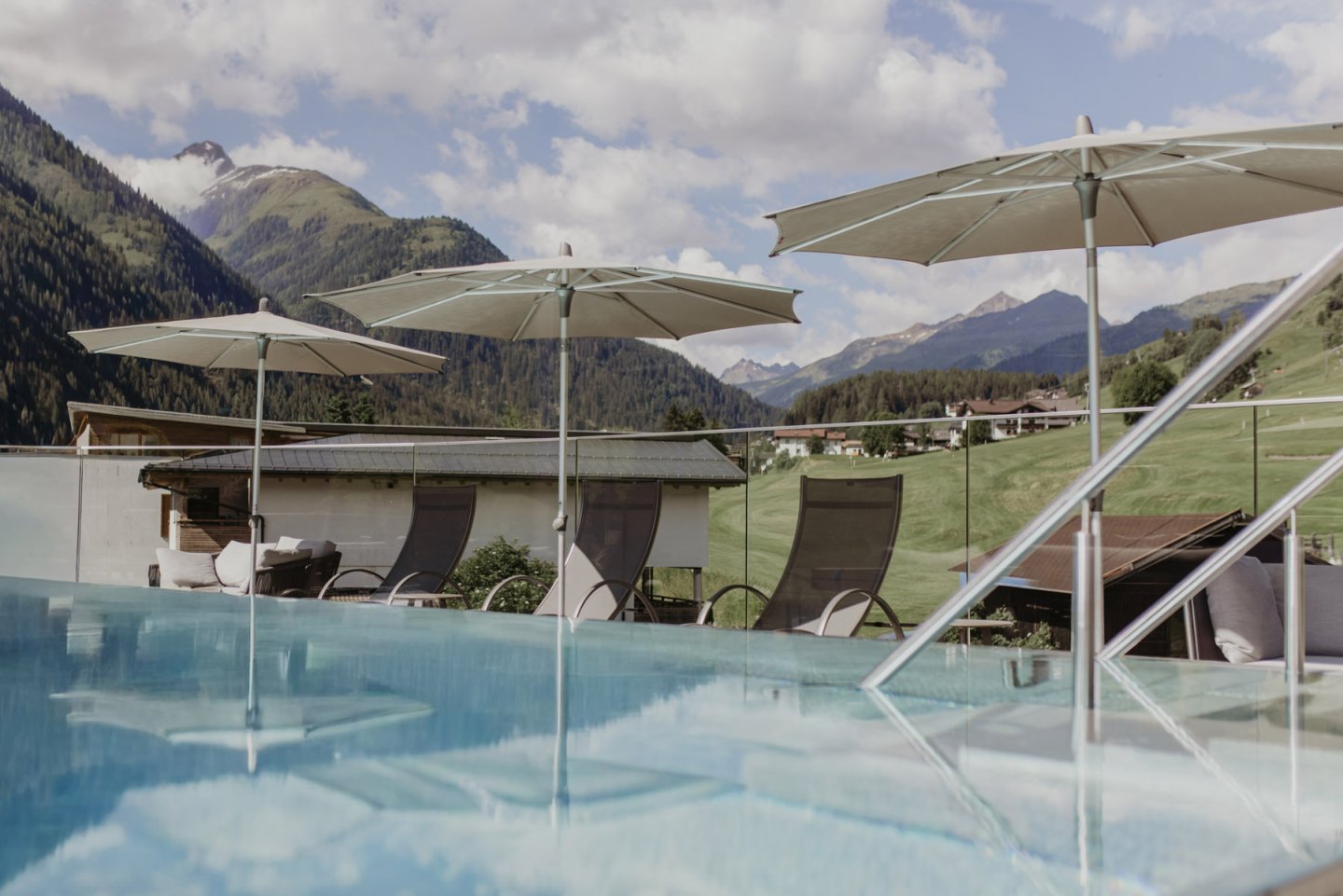 Rooftop Pool mit Sonnenschirmen und Liegen, dahinter grüne Berghänge und das Dorf