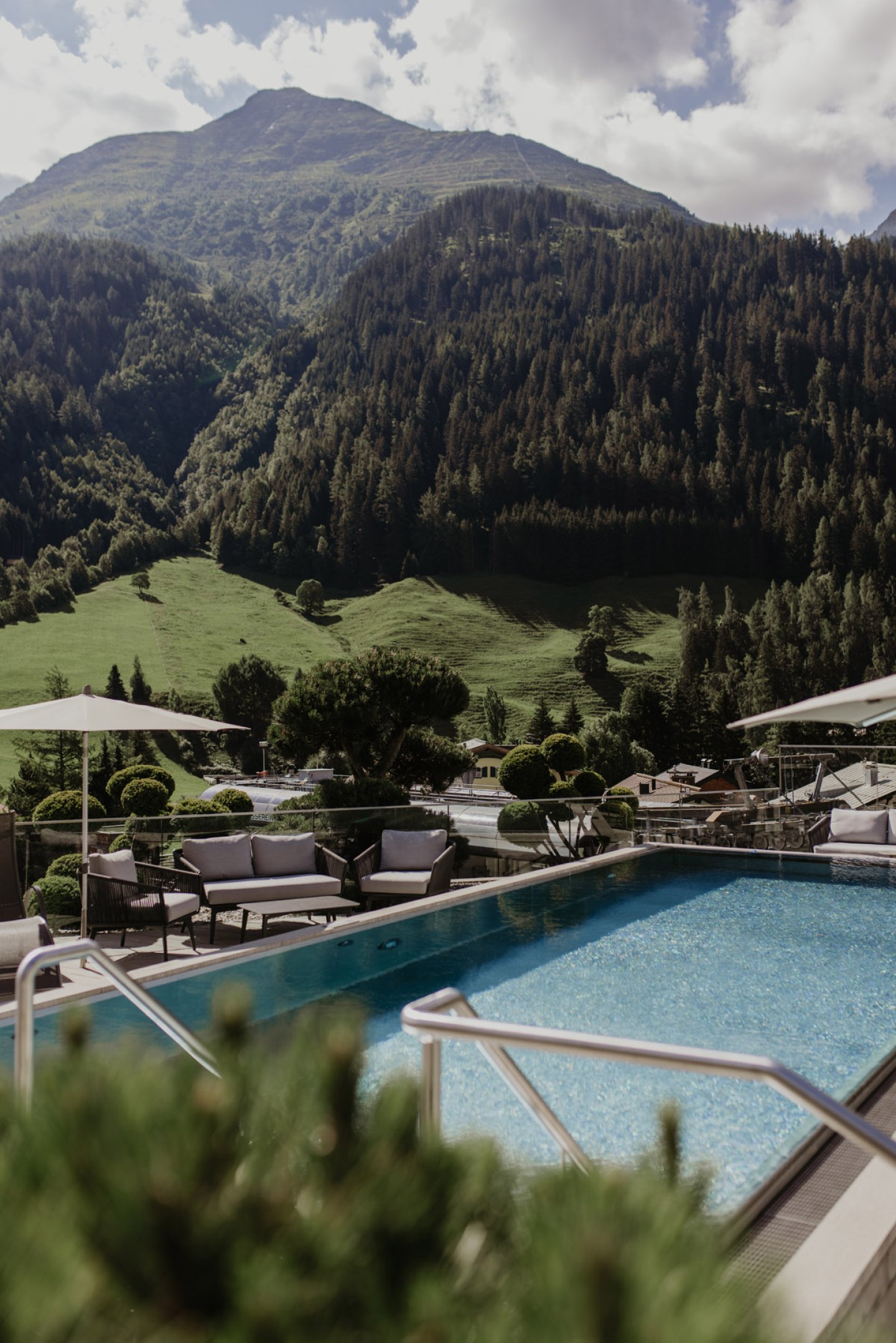 Rooftop Pool mit Sitzgruppe, dahinter ein bewaldeter Berggipfel und grüne Wiesen