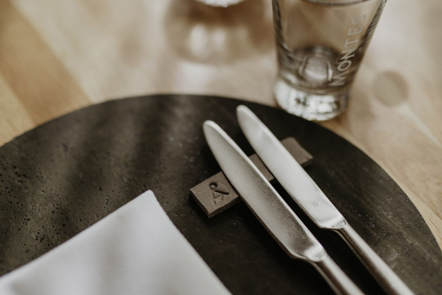 Place setting with a dark plate, white napkin and knives on a knife rest, plus a water glass